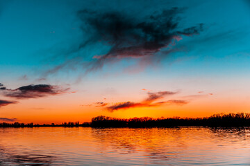 Orange, yellow, pink, blue and purple sunset sky  with dark colorful clouds with black trees reflection on calm river water	