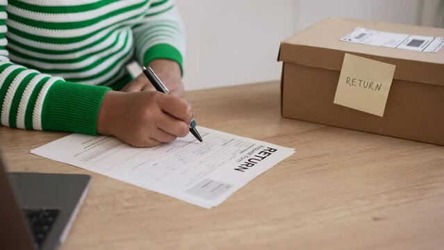 Young female client is filling out return request form and sitting at desk in home room spbd. 4k Close view of american african woman writes on paper with pen and wants to return parcel back, uses