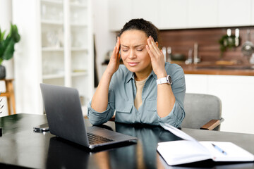 Frustrated young woman using laptop computer suffering from headache, sitting in home office and...