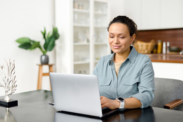 Confident lady checking emails, conducting business correspondence while sitting at the table, looking at the screen. Smiling business woman using laptop for remote work from home