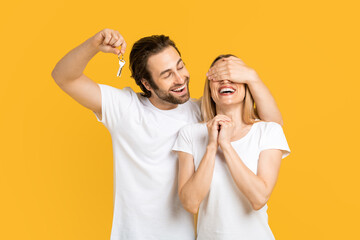 Smiling young european guy in white t-shirt closes eyes to his wife and holds keys, isolated on yellow background