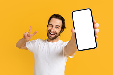 Cheerful emotional young european guy in white t-shirt show phone with empty screen and peace sign