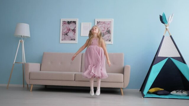 Cute little girl with long blond hair in lilac dress laughs merrily and jumps near sofa, 4k: Gomel, Belarus - Febr 2021