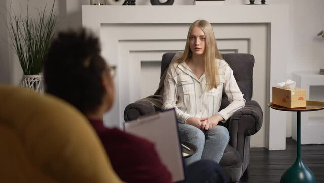 Young female client is talking and psychologist listening while sitting on couch in interior spbas. 4k Attractive blonde speaks and looks with smile, American African woman listens to her and holds