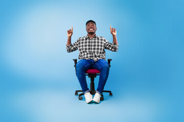 Fototapeta na wymiar Attractive young black guy pointing up at copy space, sitting on chair over blue background, mockup for advertisement