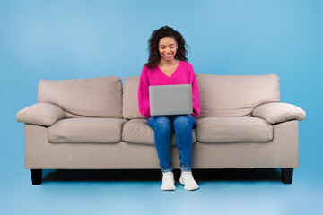 Happy young black woman in casual wear working online, sitting on sofa and using laptop on blue studio background