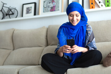 Smiling young Muslim woman using a smart phone at home. Space for text.