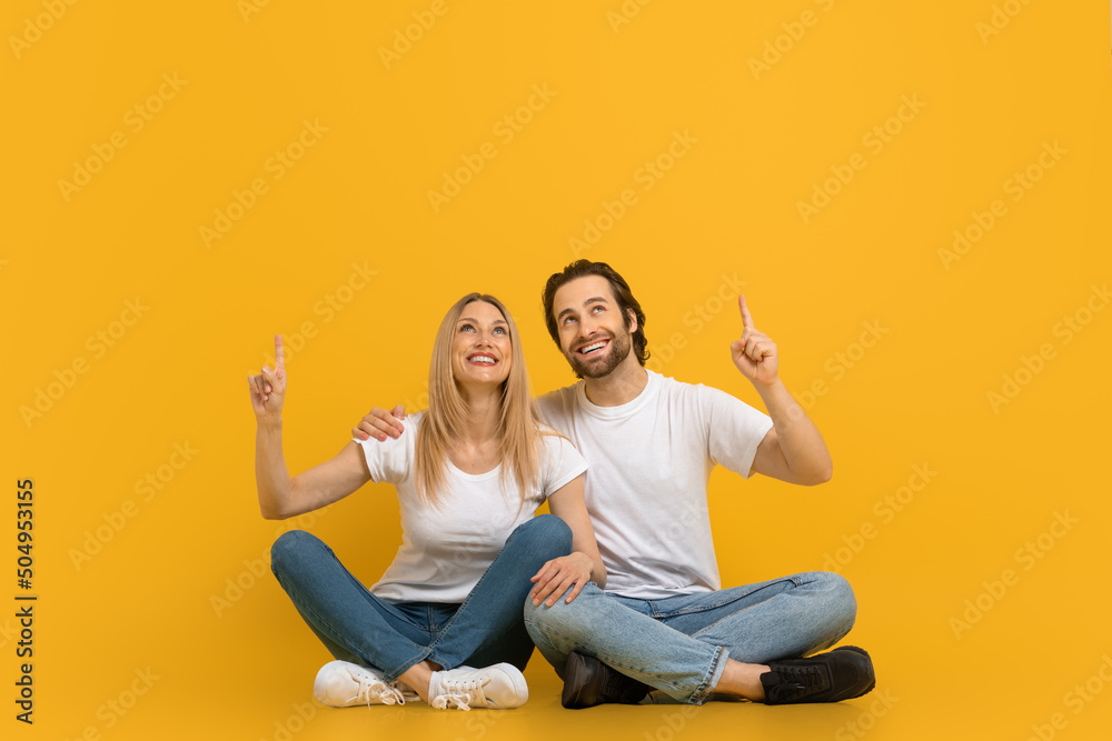 Wall mural cheerful millennial european male with beard and female hugging, sit on floor, show fingers up at fr