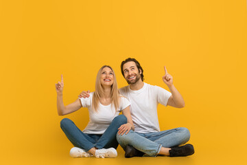 Cheerful millennial european male with beard and female hugging, sit on floor, show fingers up at free space
