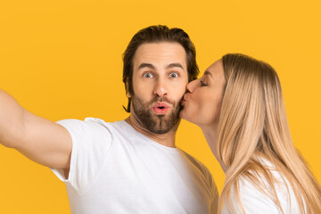 Happy young european blonde wife in white t-shirt kisses surprised husband on cheek, isolated on yellow background