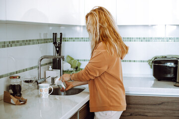 Blonde young woman in home clothes wash mugs in sink under running water after eating and tidy up...