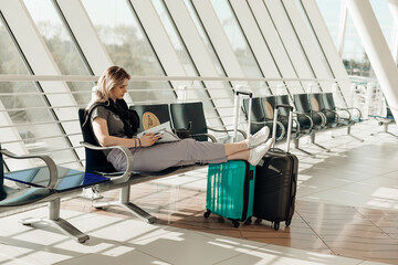 Passenger sitting on chair in modern light airport with outstretched legs on suitcases, reading...