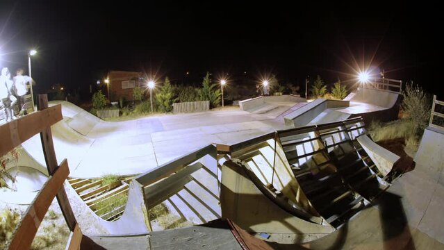 static timelapse of a wooden skatepark during the night where we can see the lights of the boys who go with the skateboard passing over the ramp and the sky with stars illuminated a wide angle image