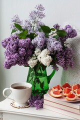 A fragrant bouquet of colorful lilacs stands in a green glass vase on a white background. Next to a cup of hot tea and sliced ​​grapefruit.