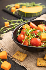 Vegetarian vegetable salad of tomatoes, pumpkin, microgreen pea sprouts on black concrete background. Side view, selective focus.