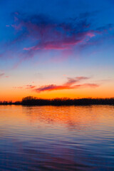 Orange, yellow and purple sunset on river with dark colorful clouds in sky with trees reflection in water