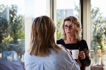 Senior ladies talking by the window. Portrait of wavy-haired woman in glasses with coffee talking...