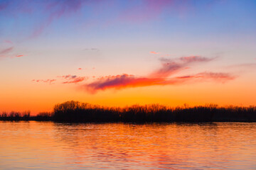 Fototapeta na wymiar Orange, yellow and purple sunset on river with dark colorful clouds in sky with trees reflection in water