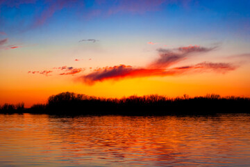 Fototapeta premium Orange, yellow and purple sunset on river with dark colorful clouds in sky with trees reflection in water