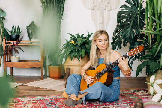 Woman sitting on a multi-colored carpet against the background of plants plays the acoustic guitar
