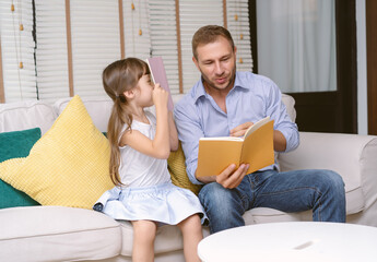 Handsome father sitting on sofa reading a story book to his daughter in the living room at home....