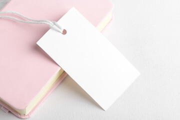 Rectangle white gift tag mockup with white cord, close up on pink book and white background. Blank...
