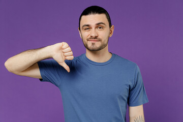 Obraz premium Young displeased dissatisfied sad disappointed man 20s wear basic blue t-shirt showing thumb down dislike gesture isolated on plain purple color background studio portrait. People lifestyle concept