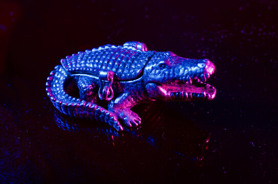 A Close up of a Small Metal  Reptilian Crocodile Jewelry Lock Box with curious synth-wave high contrast bright blue and pink night lighting on a black background.