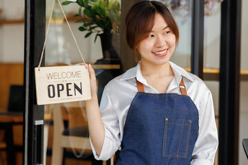 Portrait of a positive Asian businesswoman standing at the cafeteria door entrance. A cheerful young waitress in a blue apron near a glass door with an open signboard.SME Excited small business owner.