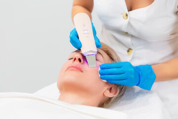 A professional cosmetologist in blue medical gloves does an ultrasound facial cleansing for an adult Caucasian woman. Cleansing device on the client's cheek. Hardware anti-aging cosmetology