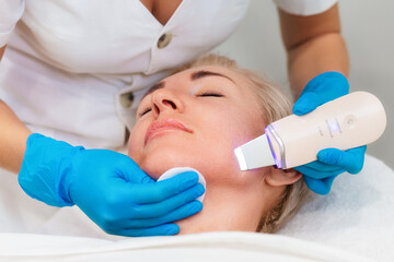 Hardware professional cosmetology. A beautician in blue medical gloves does an ultrasound facial cleansing for an adult Caucasian pretty woman. Cleansing device on the client's chin