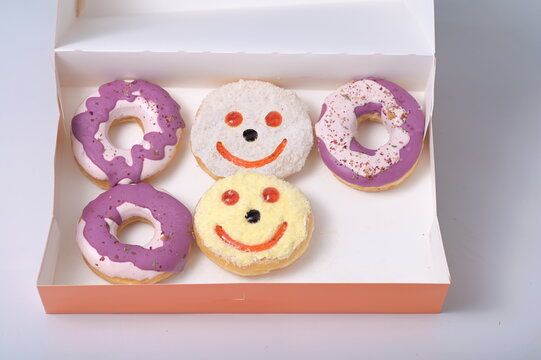 Five donuts placed in an orange cardboard box. 2 donuts coated in purple sugar, 2 painted with smiles and eyes with red jam, one with white coated. Missing one delicious donut. is a popular dessert
