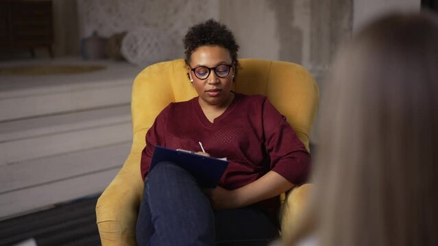 Female psychologist talking to client and writing on clipboard, sitting on couch in room spbas. 4k Closeup view of beautiful african american woman listens to story and talks with smile, takes notes