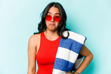 Young hispanic woman holding beach towel isolated on blue background shrugs shoulders and open eyes...