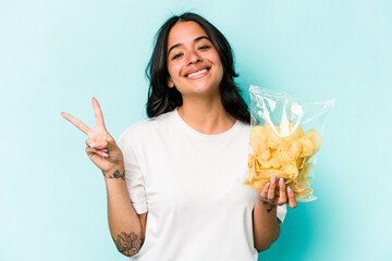 Young hispanic woman holding a bag of chips isolated on blue background joyful and carefree showing...