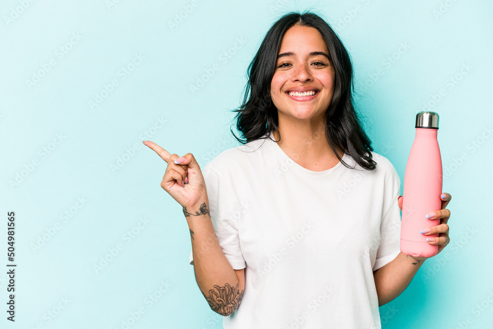 Wall mural young hispanic woman holding thermo isolated on blue background smiling and pointing aside, showing 