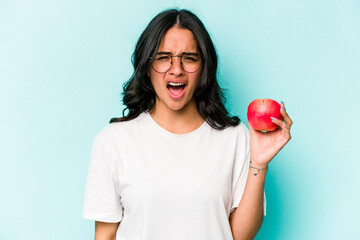 Young hispanic woman holding an apple isolated on blue background screaming very angry and...
