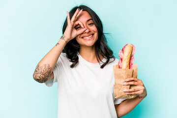 Young hispanic woman eating a sandwich isolated on blue background excited keeping ok gesture on eye.