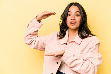 Young hispanic woman isolated on yellow background shocked and amazed holding a copy space between hands.