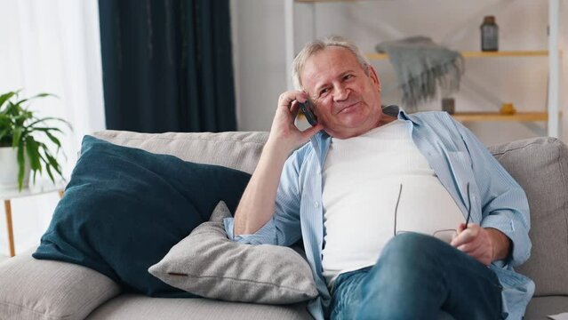 Cheerful old man talking by phone while sitting on the sofa at home
