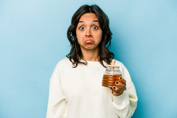 Young hispanic woman holding a honey isolated on blue background shrugs shoulders and open eyes confused.