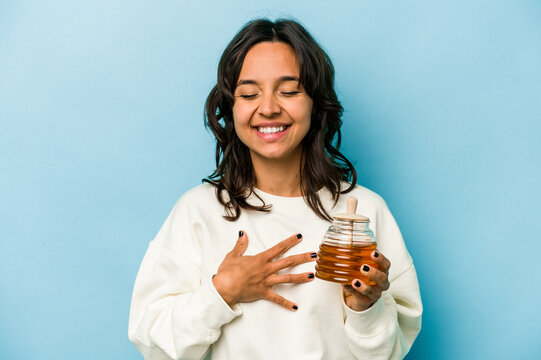 Young hispanic woman holding a honey isolated on blue background laughs out loudly keeping hand on chest.