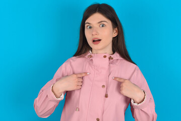 Embarrassed young beautiful Caucasian woman wearing pink raincoat  indicates at herself with puzzled expression, being shocked to be chosen to participate in competition, hesitates about something