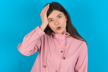 Embarrassed young beautiful Caucasian woman wearing pink raincoat over blue wall with shocked...