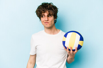 Young caucasian man playing volleyball isolated on blue background confused, feels doubtful and...