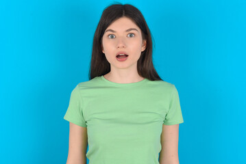 Shocked young beautiful Caucasian woman wearing green T-shirt over blue wall stares bugged eyes keeps mouth opened has surprised expression. Omg concept