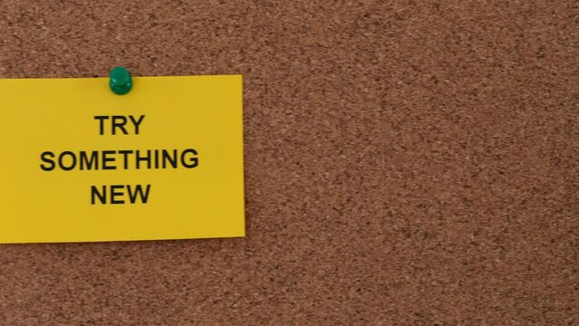 A sticky note with the words Try Something New on it pinned to a cork board. Close up.