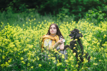 A cute European girl with long hair with her pet black poodle in a meadow with yellow flowers. Walking with a dog in nature