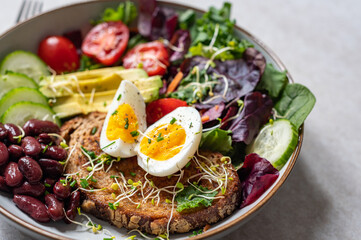 Light green salad with leaves, avocado, cherry tomatoes, cucumber slices, red beans, micro greens and medium boiled eggs on slice of toasted bread.