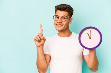 Young caucasian man holding a clock isolated on blue background showing number one with finger.